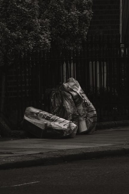 A narrow urban alleyway cluttered with a large, sealed black rubbish bag in the foreground, positioned on the dirty pavement. Behind it, there is an accumulation of discarded cardboard boxes, plastic 