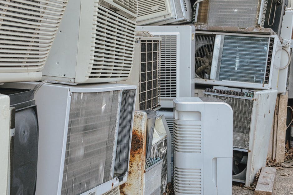 A collection of various discarded air conditioning units and external cooling appliances stacked outdoors on a paved surface, primarily consisting of white, grey, and metallic finishes with some rust 